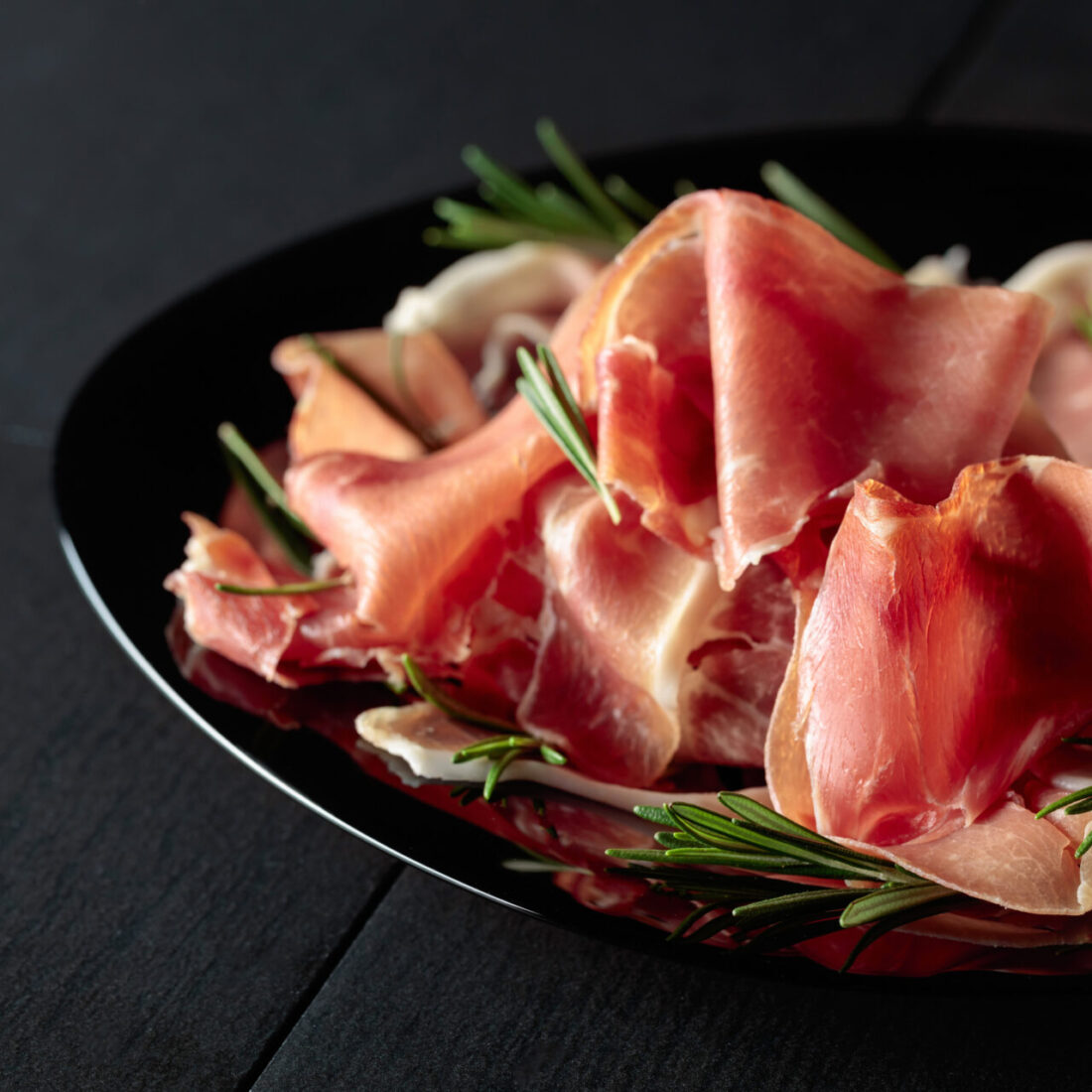 Italian prosciutto or Spanish jamon with rosemary on a black plate.